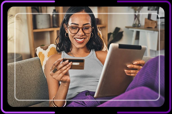 Imagem de mulher no sofá segurando cartão de crédito com uma mão e um tablet com outra