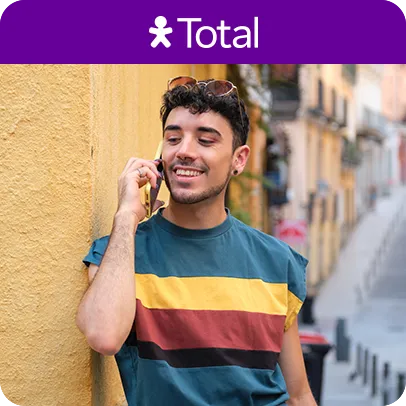Homem com camiseta listrada, encostado em parede amarela, falando ao telefone.