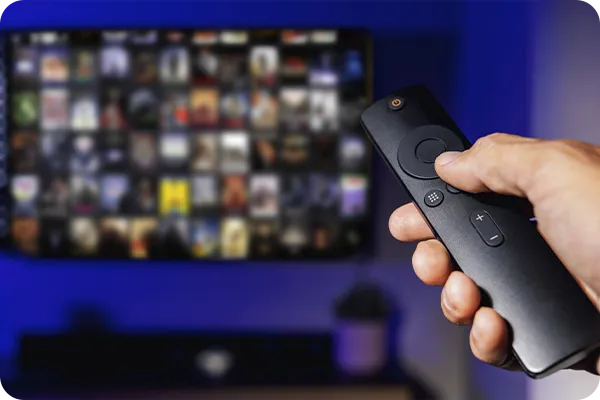 Mão segurando controle remoto em frente a uma TV com catálogo de streaming.