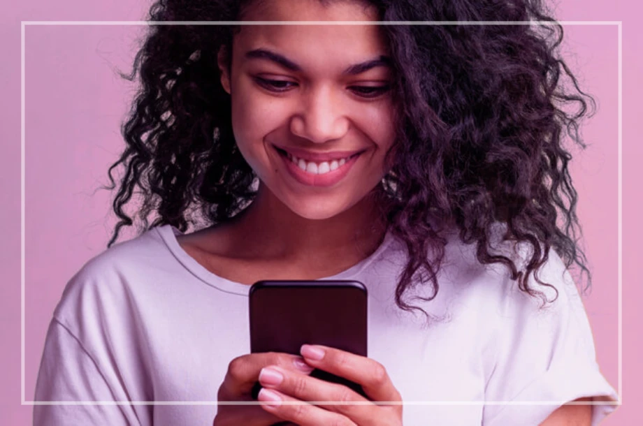 Mulher sorrindo usando celular com fundo rosa.