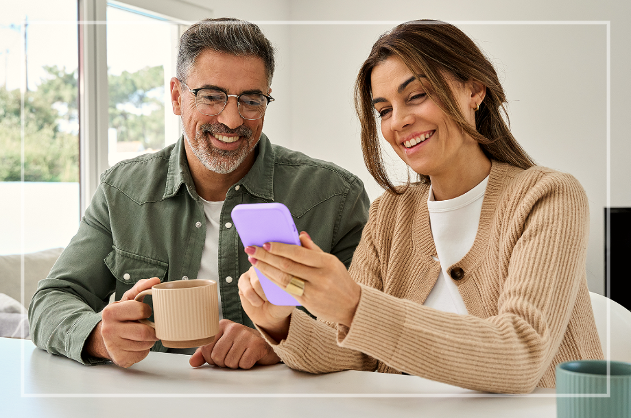 Casal sorrindo olhando um celular juntos enquanto o homem toma café.