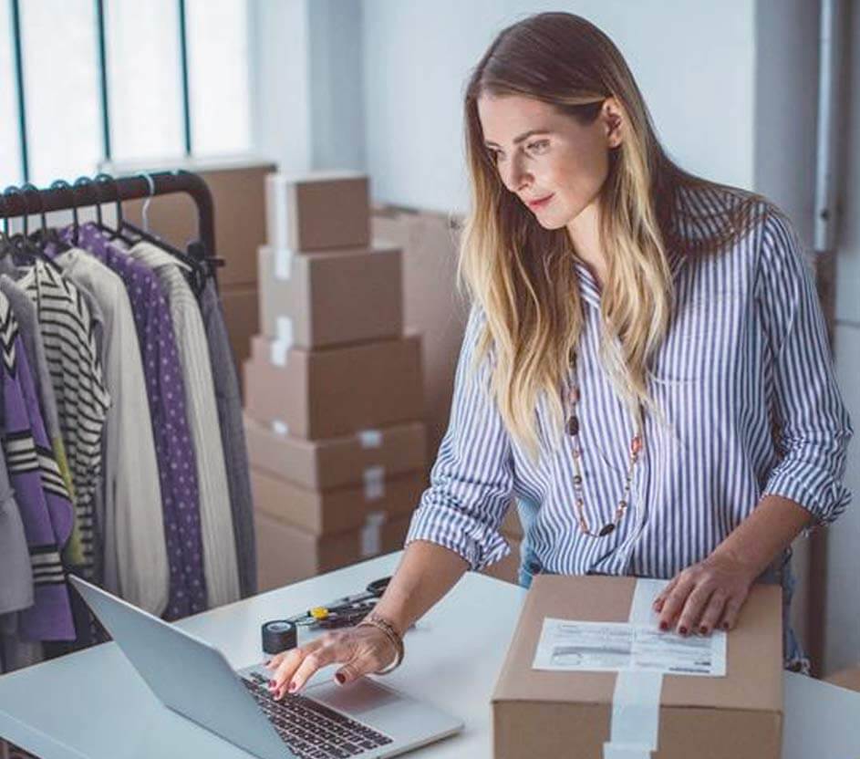 Imagem de uma mulher que, em uma das mãos utiliza um notebook, e a outra sobre uma caixa. 