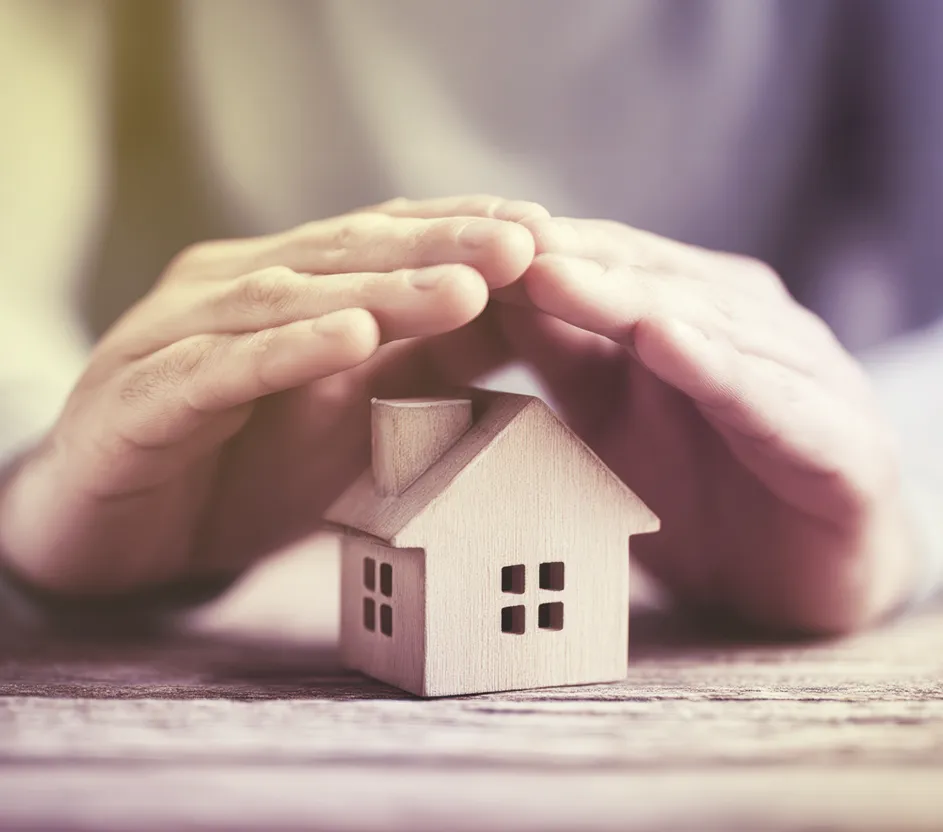 Mãos cobrindo uma casa de madeira em miniatura.