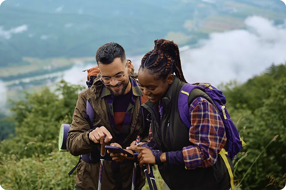 Casal com equipamentos de trilha olhando para smartphone, ao fundo paisagem de floresta.