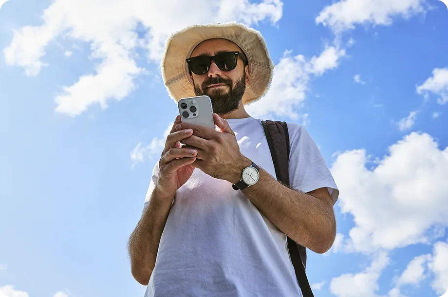 Homem com chapéu e celular na mão, ao fundo céu azul com nuvens.