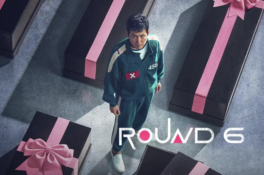 Imagem do personagem 456 da série Round 6, em pé olhando para a câmera ao lado de caixões pretos.