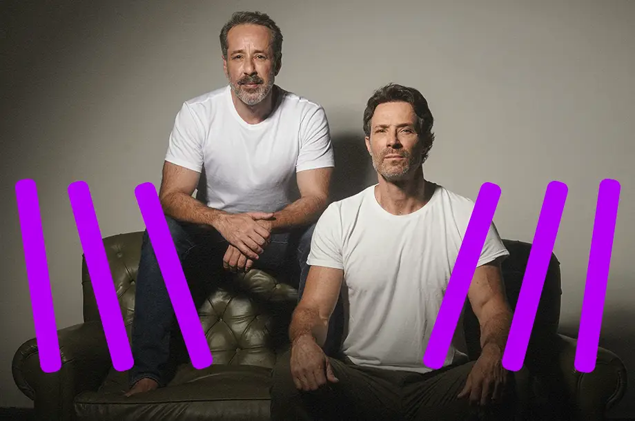 Dois homens de camiseta branca sentados em um sofá olhando para frente com uma cara séria.