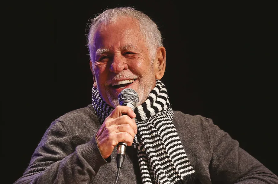 Nelson Motta com cachecol listrado, sorrindo e segurando um microfone.