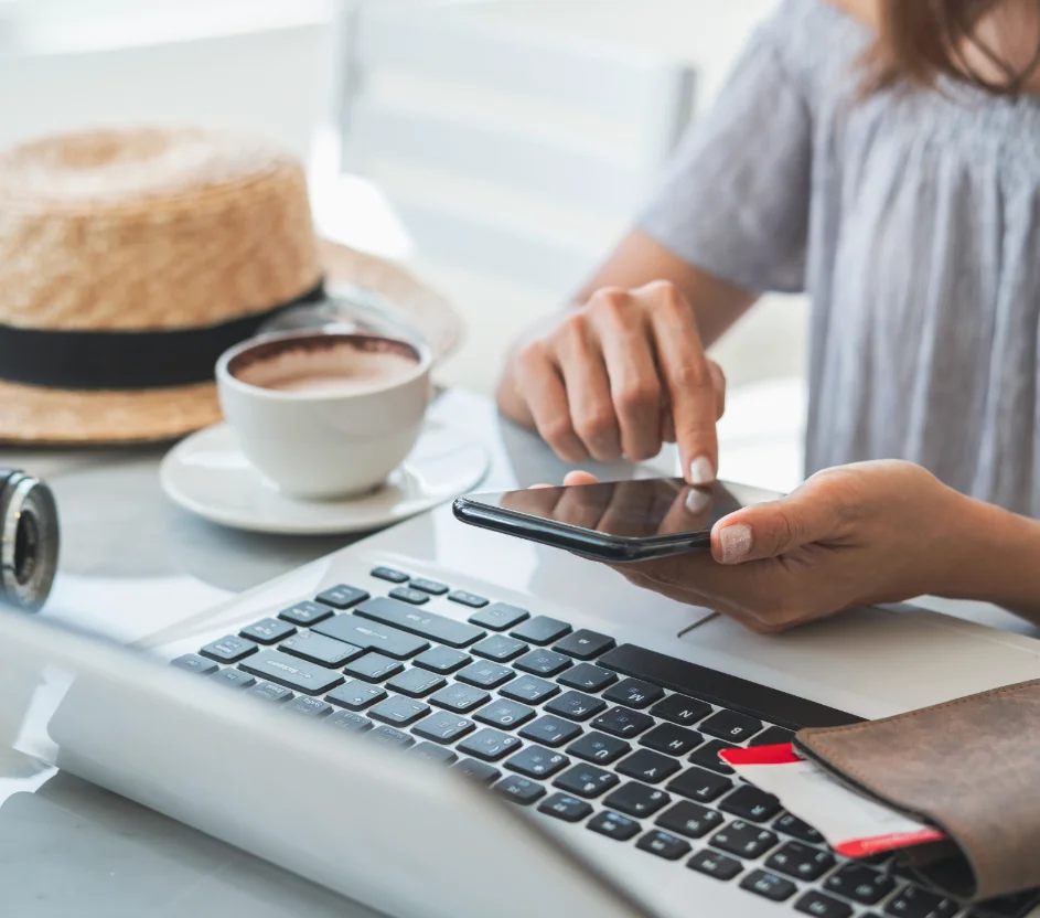 Mulher sentada na mesa com celular, notebook, câmera fotográfica, xicara de café e chapéu na mesa. 