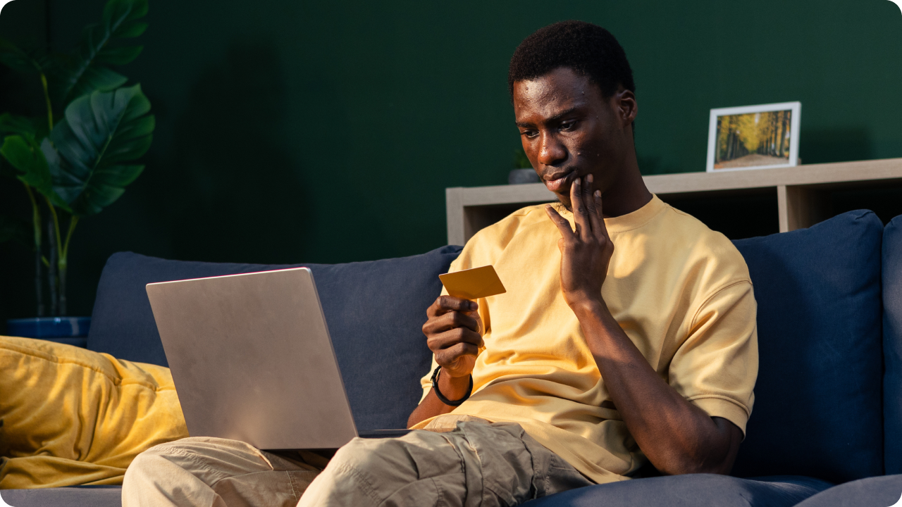 Homem negro  sentado em sofá com a mão no rosto e segurando um cartão de crédito diante de notebook.