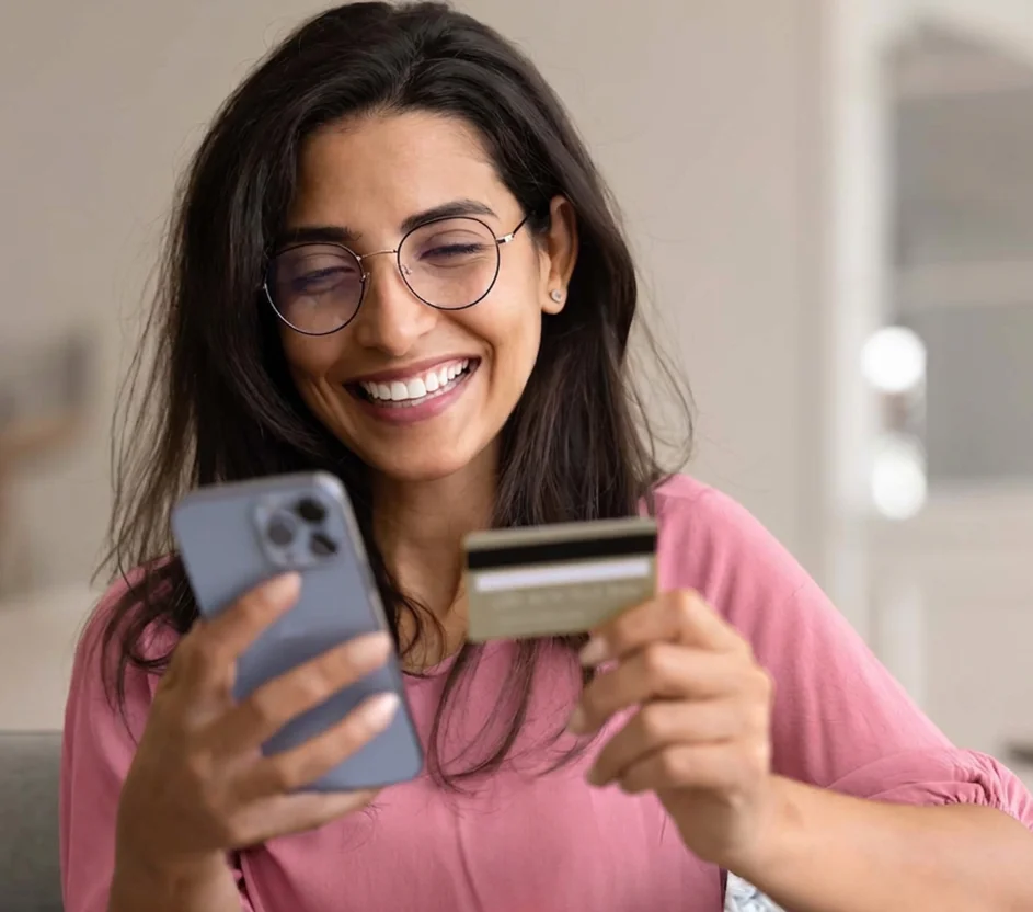 Mulher branca segura cartão bancário enquanto utiliza smartphone para comprar pela internet.