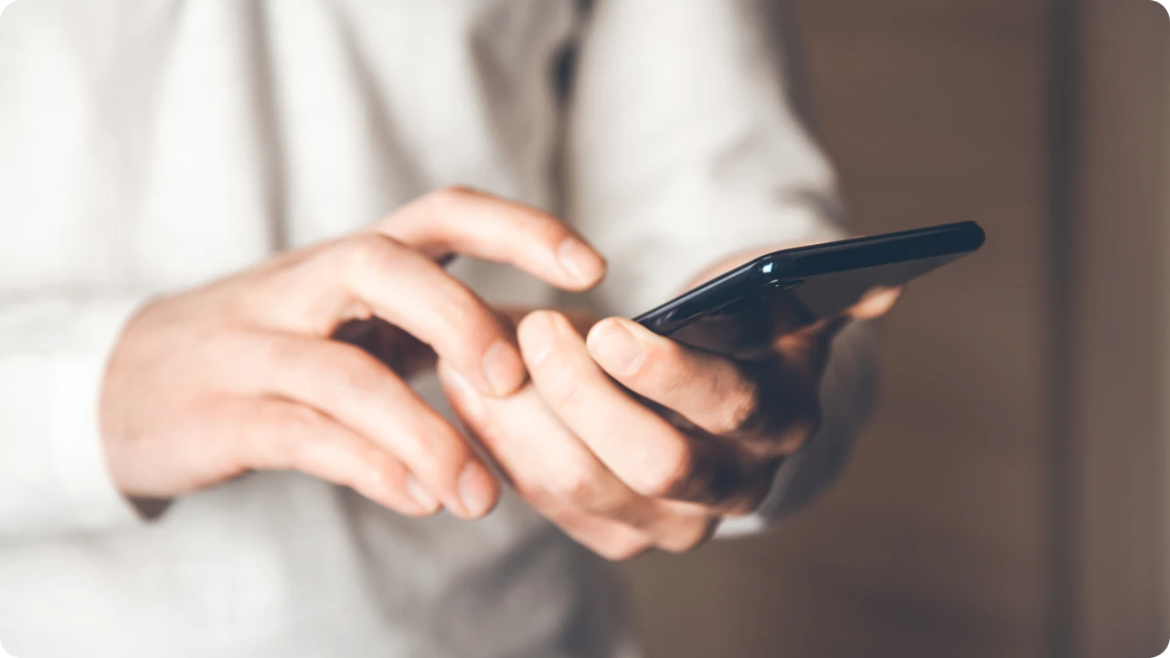 Mãos interagem com celular em destaque sobre fundo desfocado.