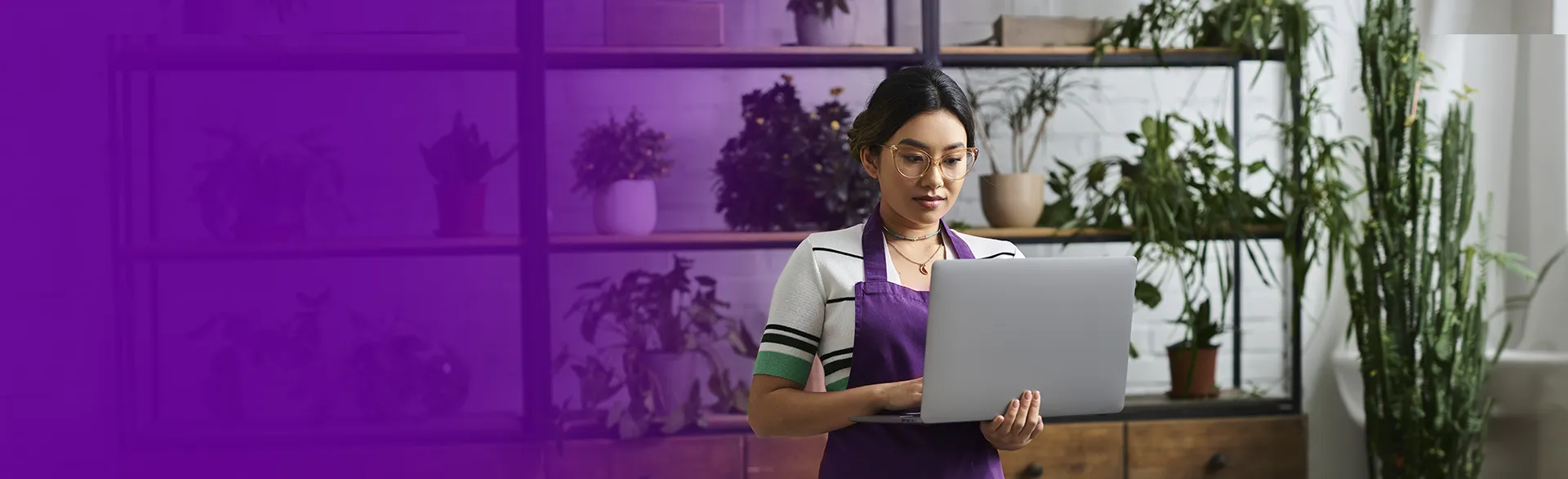 Mulher com avental roxo segurando notebook, ao fundo prateleira com plantas.