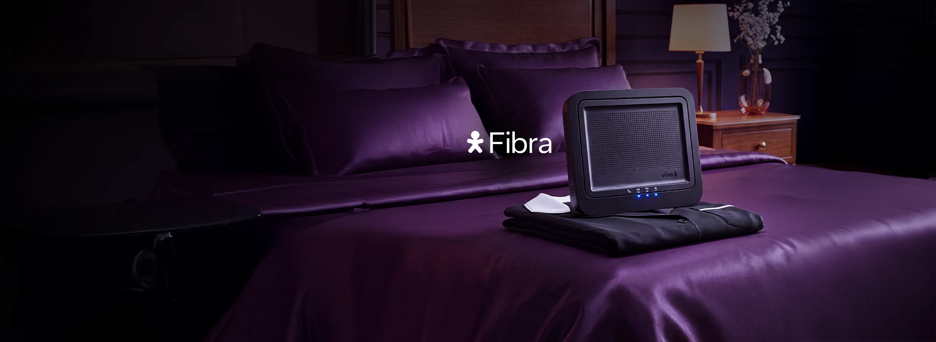 Um quarto com a cama de lençóis roxo e um moldem de Fibra em cima de um uniforme. Logo Vivo Fibra.