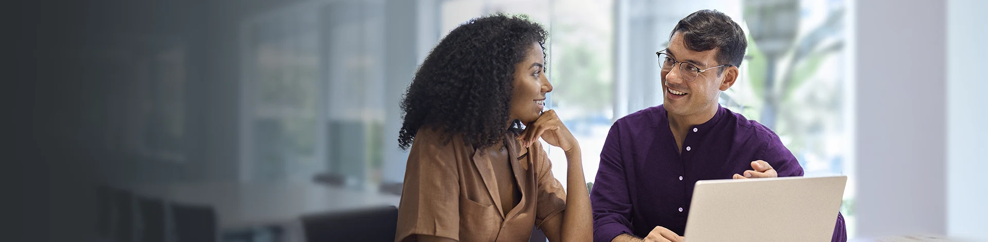 Uma mulher conversando com um homem ao seu lado, os dois sentados em uma mesa com um notebook.