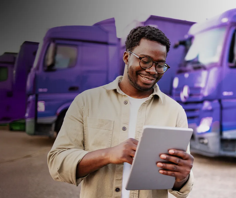 Imagem de um homem sorrindo mexendo em um tablet e atrás caminhões estacionados