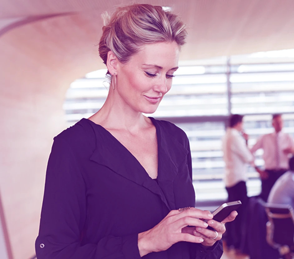 Mulher loira de cabelos presos com celular na mão olhando pra ele em um ambiente corporativo.