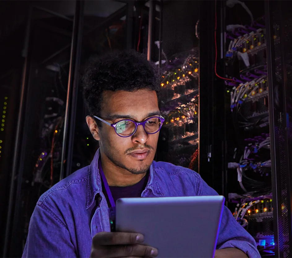 Homem de óculos com um tablet na mão em uma sala de data center.
