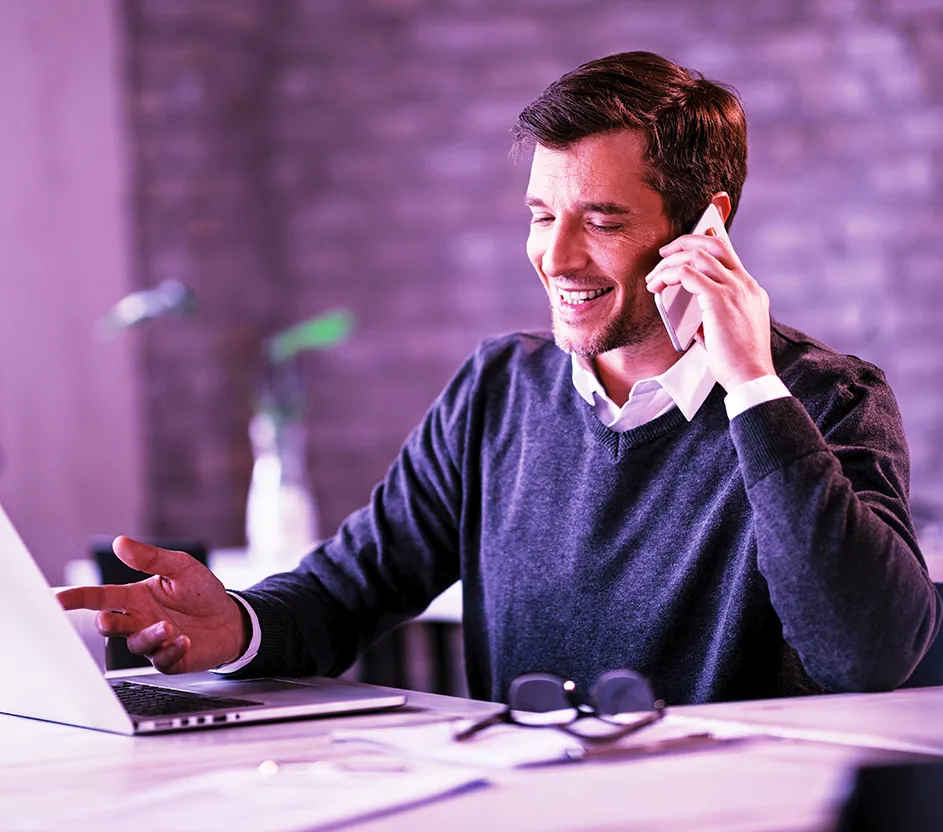 Homem sorrindo falando no celular e usando notebook em um ambiente de escritório.