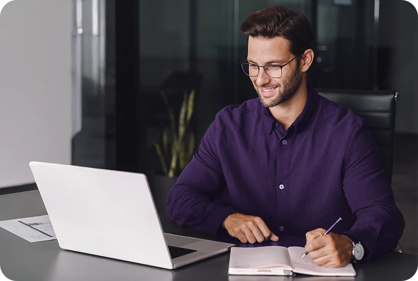 Pessoa sentada em escritório acompanhando conteúdo no notebook e anotando informações.