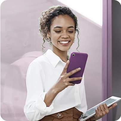 Imagem de pessoa segurando celular com uma mão e tablet com a outra, ela está encostada em um vidro