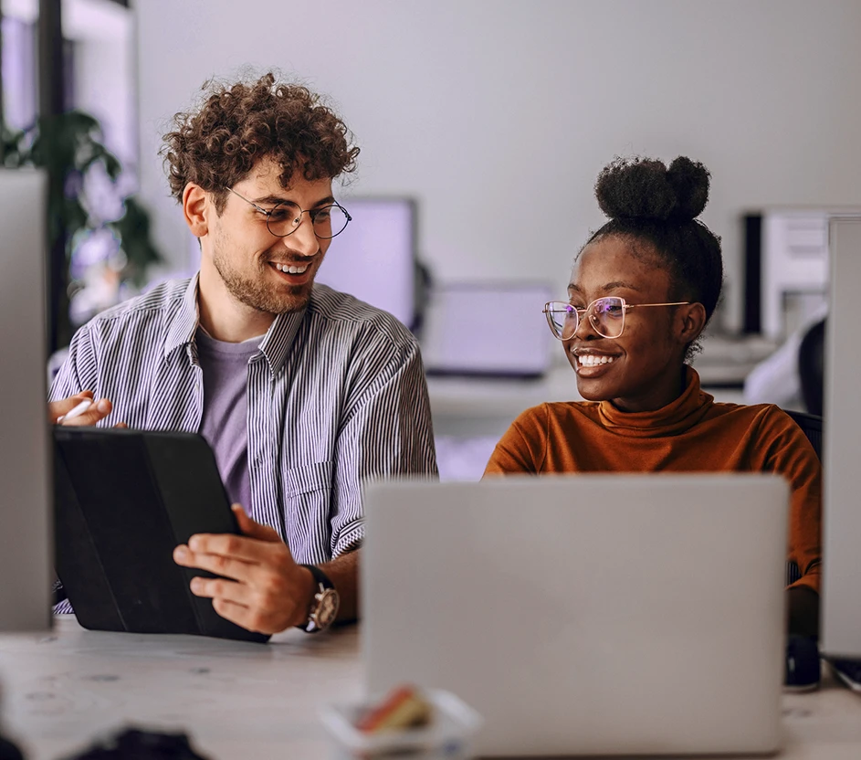 Imagem de um homem segurando um tablet enquanto conversa com uma mulher em frente ao notebook.