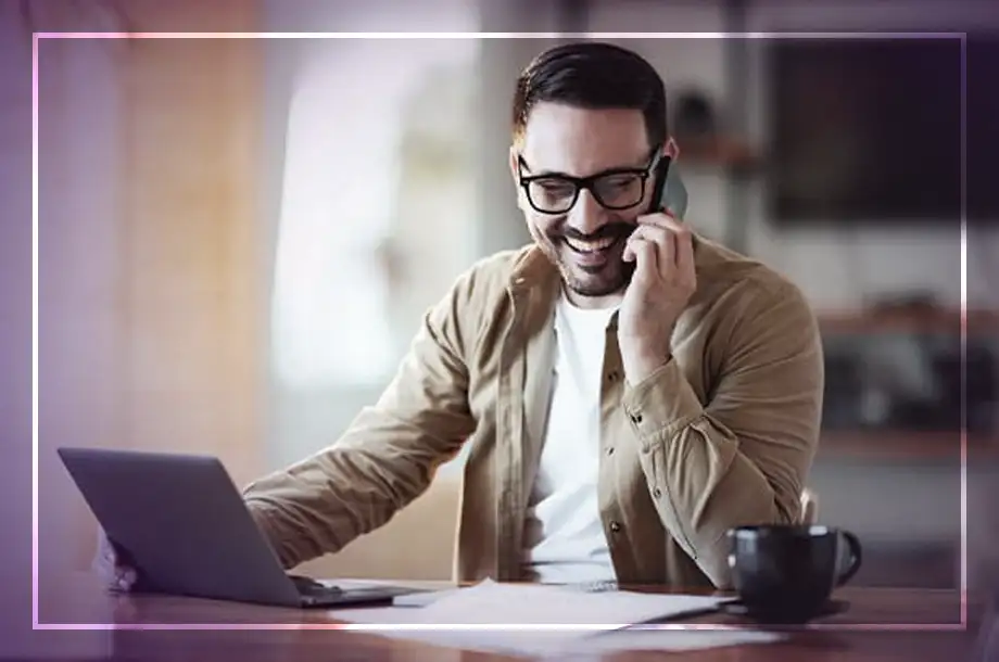 Pessoa em mesa com notebook e celular. Caneca ao lado. Fundo desfocado.