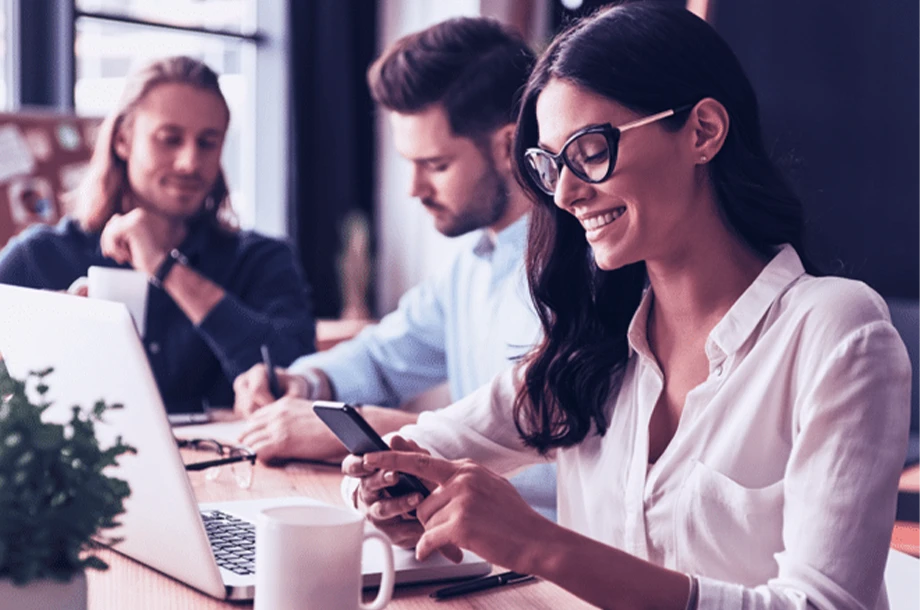 Colegas de trabalho sentados um ao lado do outro com destaque em uma mulher no celular e notebook.
