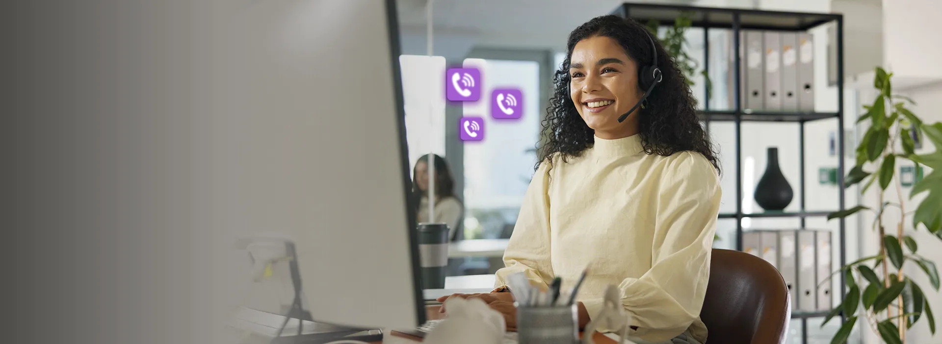 Imagem de uma mulher sorrindo com fone de ouvido e microfone olhando para uma tele de computador