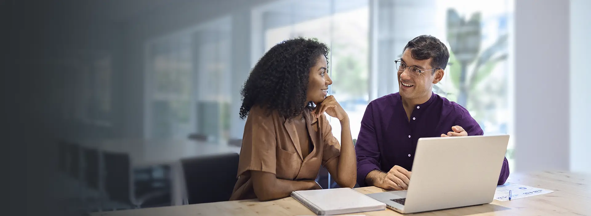Duas pessoas conversando em uma mesa com um notebook em cima em um ambiente de escritório.