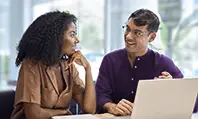 Duas pessoas conversando em uma mesa com um notebook em cima em um ambiente de escritório.